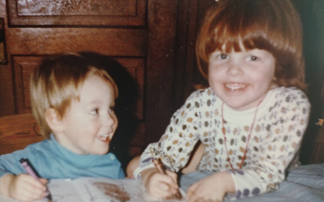 A young boy in a blue shirt looks at a young girl with red hair as they both smile, and hold crayons to a colouring book.